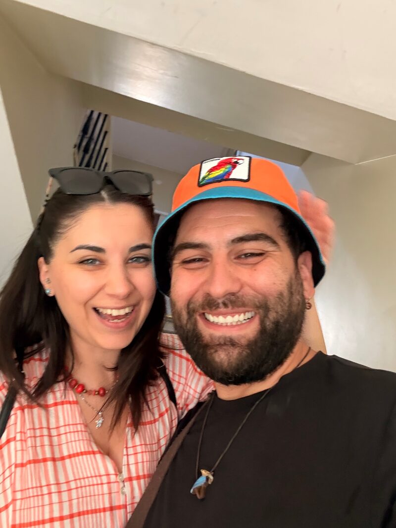 Bir merdiven boşluğunda çekilmiş, biraz bulanık bir selfie. Solda Seda: uzun koyu saçlı, güneş gözlüğü saçlarının üstünde; kırmızı-beyaz kareli bir gömlek, kırmızı boncuklu bir kolye ve altında küçük uçlu ince gümüş bir zincir, kulağında küçük turkuaz bir küpe; ağzını kocaman açmış, kahkahayla gülüyor. Sağda Tuncay: koyu, gür sakallı; başında turkuaz bantlı, altı siyah kenarlı turuncu bir balıkçı şapkası, önünde rengârenk bir papağan arması; siyah bir tişört, kulağında küçük bir küpe, boynunda koyu bir ipe takılı mavi-kahverengi bir kolye ucu; ağzı kulaklarında. İkisi birbirine sokulmuş duruyor; Tuncay'ın başının arkasında havaya kalkmış, bulanık bir el görünüyor. Üstlerinde merdivenlerin eğimli beyaz alt yüzleri ve bir parça korkuluk.